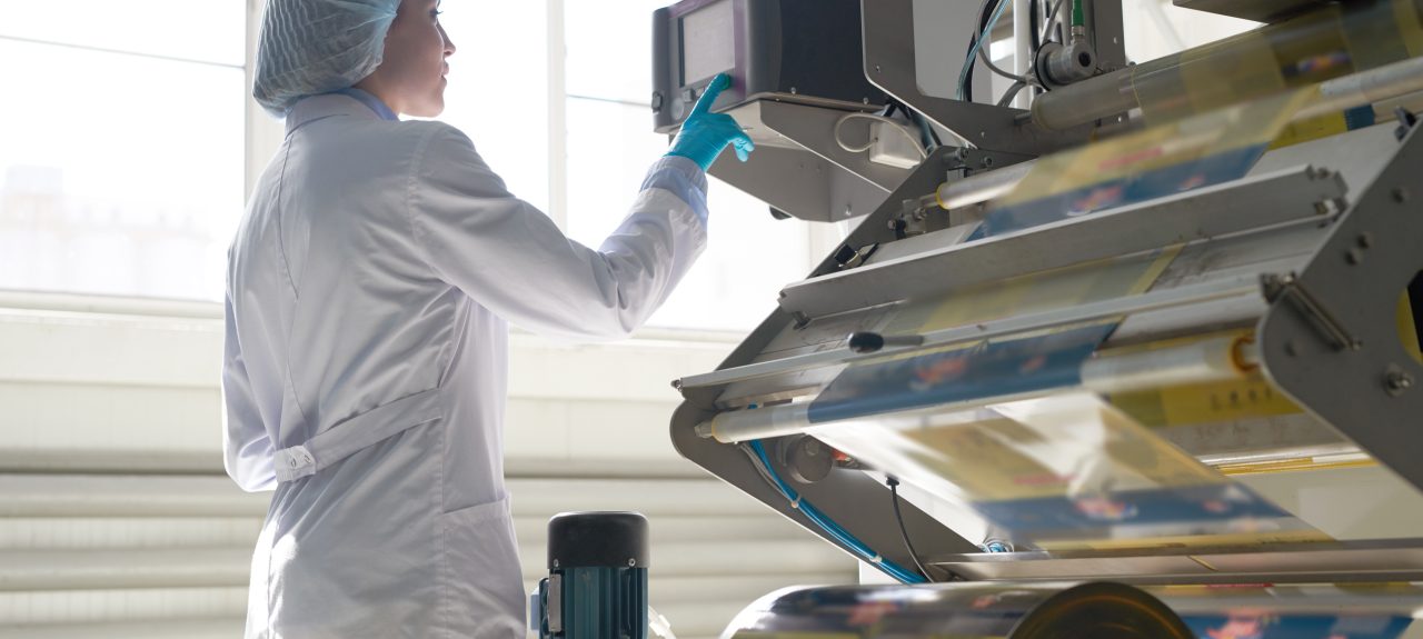 Side view portrait of female factory worker wearing clean white coat standing by machine units at packaging line, copy space